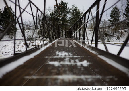 Metal Bridge Over River. The snow-covered metal bridge leads to towering pine trees, creating a serene and picturesque winter landscape that invites a quiet and peaceful retreat. 122466295