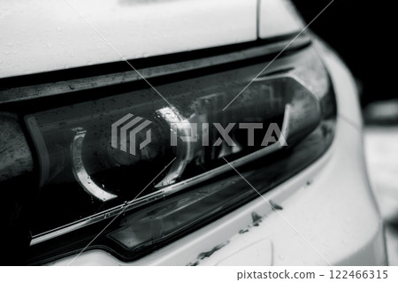 Close-up of a car headlight covered in water droplets. Close-up of a wet headlight of a modern car reflecting light in the dark, showing intricate details and design elements with water droplets. 122466315