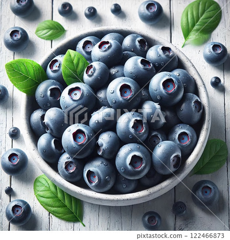 A bowl of fresh, plump blueberries with green leaves on a white wooden surface, showcasing their deep blue color and natural sheen 122466873