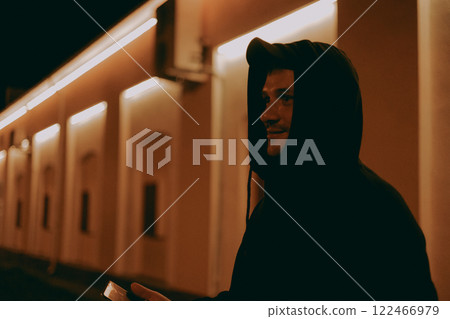 A man in a hood stands on a city street at night with a phone in his hand. The man looks to the side in the light of the city signs at night. Concept of a handsome man on a city street at night. 122466979