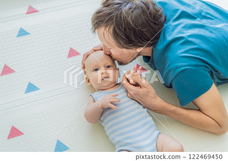 A baby lies on a soft children play mat while their father hands gently support and protect them. Heartwarming moment of care and connection. Parenthood, safety, and love concept A baby lies on a soft children play mat while their father hands gently support and protect them. Heartwarming moment of care and connection. Parenthood, safety, and love concept 122469450
