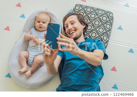 A 3-month-old baby lies safely in a cozy infant cocoon, secured with a special safety belt, while their father lies beside them during a video call. Modern parenting, bonding, and technology concept 122469466