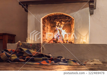 A warm and inviting scene featuring a lit fireplace with glowing firewood and candles. A colorful traditional rug and pillow lie on the floor, creating a cozy atmosphere. Home, relaxation, and rustic A warm and inviting scene featuring a lit fireplace with glowing firewood and candles. A colorful traditional rug and pillow lie on the floor, creating a cozy atmosphere. Home, relaxation, and rustic 122469478