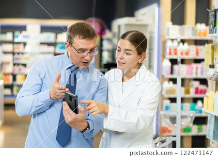 Middle-aged male customer asking young chemist about product reflected on his phone Middle-aged male customer asking young chemist about product reflected on his phone 122471147
