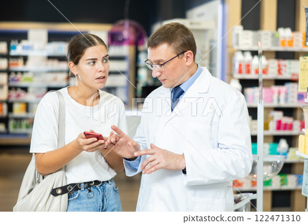 Pharmacist man communicates with girl client, looks at mobile phone screen Pharmacist man communicates with girl client, looks at mobile phone screen 122471310
