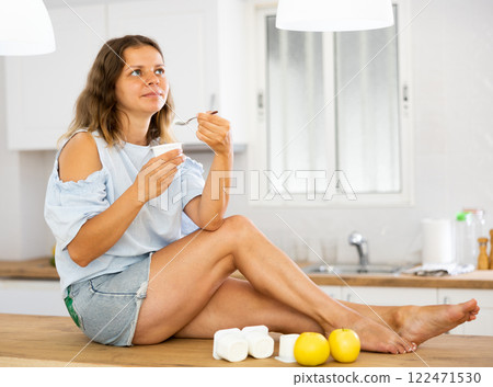 Dreamy girl eating yogurt and apples for breakfast in kitchen 122471530