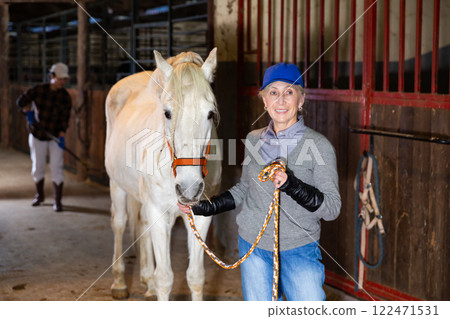 Senior woman horse breeder leading white horse through horse barn 122471531