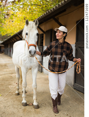 Positive woman jockey leads white horse by the bridle on the street along stable 122471699