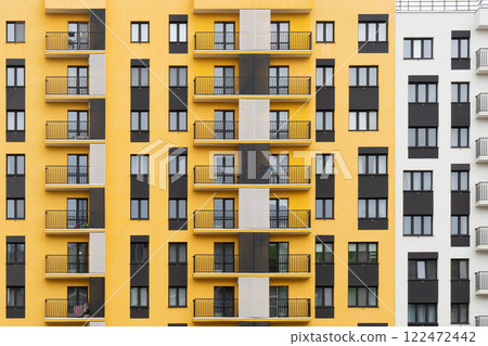 Modern apartment buildings with yellow and white facades, large windows, and balconies with metal railings, against a cloudy sky, front view Modern apartment buildings with yellow and white facades, large windows, and balconies with metal railings, against a cloudy sky, front view 122472442