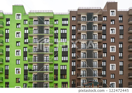 Modern apartment buildings with green and brown facades, large windows, and balconies with metal railings, standing against a cloudy sky, front view 122472445