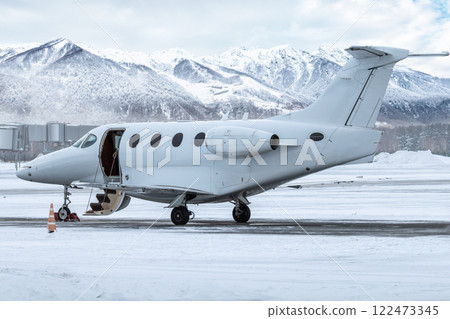 Modern white private jet with an opened gangway door at the winter airport apron on the background of high scenic snow capped mountains Modern white private jet with an opened gangway door at the winter airport apron on the background of high scenic snow capped mountains 122473345