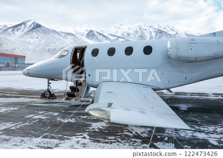 Close-up of the modern white business jet with an opened gangway door at the winter airport apron on the background of high scenic snow capped mountains 122473426