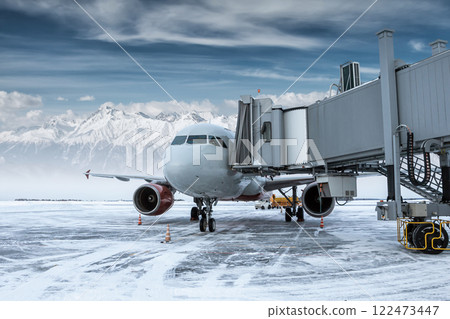 Modern passenger jetliner at the skybridge at winter airport apron on the background of high scenic mountains Modern passenger jetliner at the skybridge at winter airport apron on the background of high scenic mountains 122473447