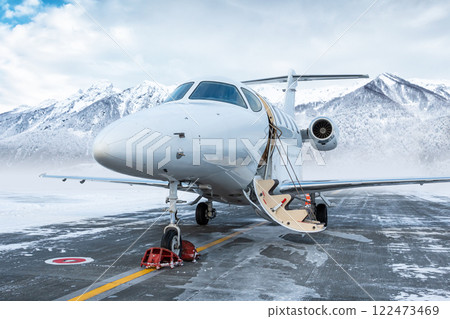 Modern white executive aircraft with an opened gangway door at the winter airport apron on the background of high scenic snow capped mountains 122473469