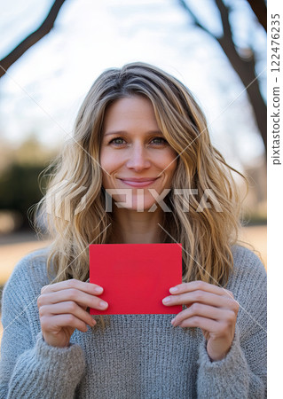 Joyful Woman Smiling While Holding Red Valentine Card in Outdoor Setting 122476235