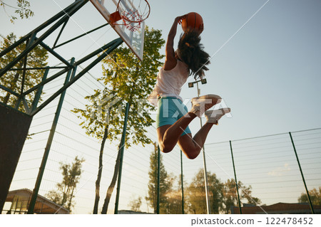 Young woman basketball player frozen in motion jumping high making slam dunk Young woman basketball player frozen in motion jumping high making slam dunk 122478452