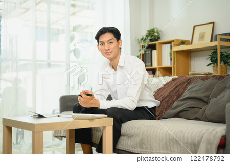 Young adult man in a white shirt sitting on couch managing his budget in a comfortable home 122478792