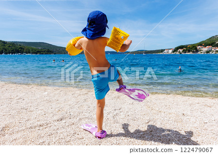 Back view cute little tanned boy kid in swimsuit sunscreen cap and fin funny walking dive in clear blue Mediterranean sea water on sunny summer day. Child have fun go snorkeling in flippers 122479067