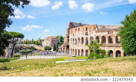 Visitors enjoy the historic Colosseum in Rome, Italy on a bright day. The ancient structure stands majestically against a backdrop of lush greenery and blue skies, showcasing its timeless beauty. 122479343