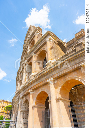 The iconic Colosseum in Rome stands tall against a bright blue sky, showcasing its ancient architecture and intricate arches. Visitors enjoy the grandeur of this historic landmark. 122479344