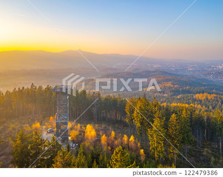 The wooden lookout tower at Cisarsky Kamen stands tall against a backdrop of vibrant autumn foliage as the sun sets, casting warm hues over the landscape near Liberec. 122479386