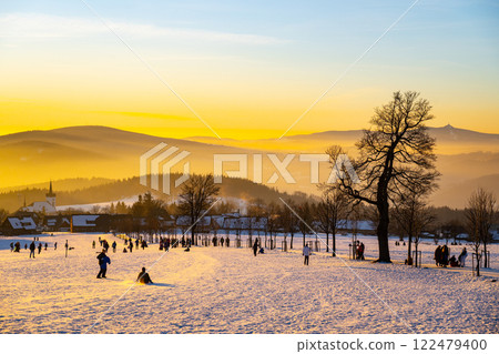 Visitors enjoy a winter sunset in Prichovice, Czechia, as they engage in various activities on the snow-covered landscape surrounded by mountains and trees. 122479400
