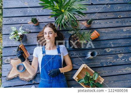An overhead shot of female gardener lying on wooden terrace holding watering can surround with potted plants, having relax time. An overhead shot of female gardener lying on wooden terrace holding watering can surround with potted plants, having relax time. 122480036