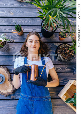 An overhead vertical portrait female gardener lying on terrace covered with potted plants, a watering can and other gardening equipment. Moment of calm in a troubled world 122480044