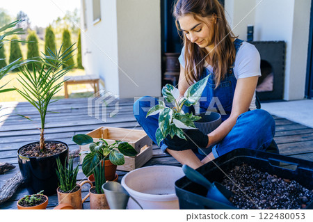 Casual lifestyle series in modern scandinavian interior. Happy home gardener transplanting plants: woman examines leaves and plant root in domestic garden outdoor on terrace of her house. Casual lifestyle series in modern scandinavian interior. Happy home gardener transplanting plants: woman examines leaves and plant root in domestic garden outdoor on terrace of her house. 122480053