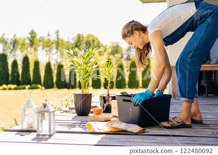 Side view woman gardener in gloves wear jeans overalls with soil, seedling, flowerpot, plants in pot preparing ground for replanting outdoor in terrace, sunny day, spring. 122480092