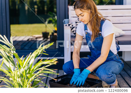 Ethnicity woman in casual clothes gardener re-potting plants on terrace at backyard outdoor. Female with gloves mixing soil in a container. Leisure, lifestyle, mental health, real life. 122480096