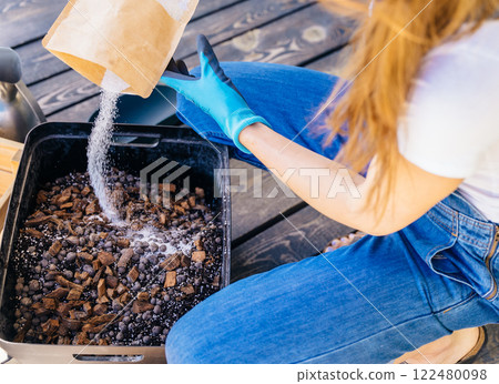 View from above of unrecognizable housewife woman in gloves and casual home clothes mix soil, preparing for plant transplantation on wooden floor terrace outdoor. 122480098