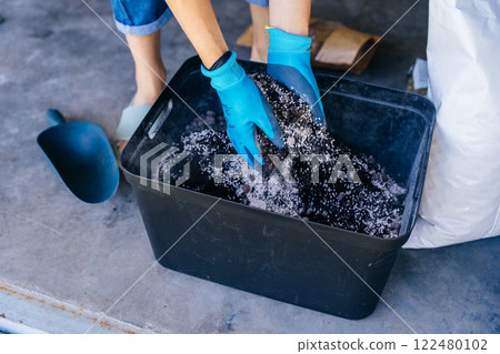 Unrecognizable housewife woman in gloves and casual home clothes mix soil, preparing for plant transplantation in garage or utility room. Close up 122480102
