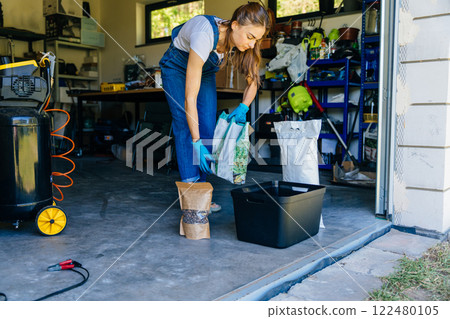Young housewife woman in casual home clothes holds a large package holds a large package. preparing soil for plant transplantation in garage or utility room. Young housewife woman in casual home clothes holds a large package holds a large package. preparing soil for plant transplantation in garage or utility room. 122480105