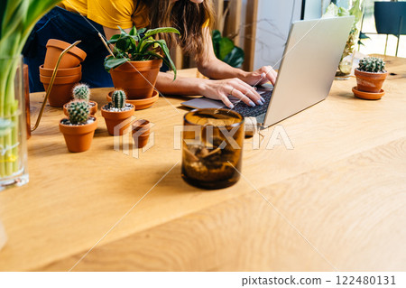 Unrecognizable young woman botanist hobbyist shopping for pots and soil for her hobby in online store at home. Close up of hands on notebook keyboard, wooden table with clay pots, plants,. 122480131