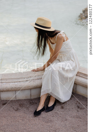 young Africa American woman in white dress straw hat sits white marble fountain and touches water  122480177