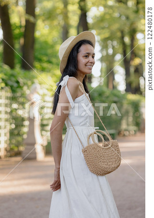 African American woman in white dress, hat walks in park poses against background of green hedge African American woman in white dress, hat walks in park poses against background of green hedge 122480178