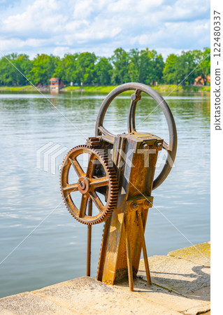 A weathered water wheel stands beside Svet Pond in Trebon, Czechia, surrounded by lush greenery. The tranquil water reflects the blue sky and clouds, creating a serene atmosphere. 122480337
