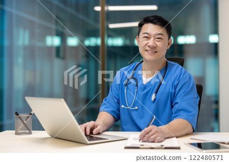 Asian doctor with stethoscope smiles confidently at desk, working on laptop in modern office. Portrays professional dedication and approachability in healthcare setting. Asian doctor with stethoscope smiles confidently at desk, working on laptop in modern office. Portrays professional dedication and approachability in healthcare setting. 122480571
