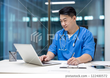 Asian man doctor in blue scrubs focused on laptop work, multitasking at desk. Uses technology to enhance patient care, exemplifying commitment to modern healthcare solutions. Asian man doctor in blue scrubs focused on laptop work, multitasking at desk. Uses technology to enhance patient care, exemplifying commitment to modern healthcare solutions. 122480572
