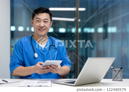 Asian man doctor holds tablet smiling in modern office with laptop, representing technology in healthcare. Blue medical uniform, stethoscope around neck, creating professional atmosphere. Asian man doctor holds tablet smiling in modern office with laptop, representing technology in healthcare. Blue medical uniform, stethoscope around neck, creating professional atmosphere. 122480579
