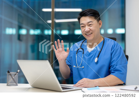 Asian doctor in blue scrubs waves and smiles at laptop, demonstrating telemedicine. Friendly and approachable, showcases modern healthcare communication and technology. Asian doctor in blue scrubs waves and smiles at laptop, demonstrating telemedicine. Friendly and approachable, showcases modern healthcare communication and technology. 122480592