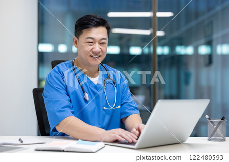Asian man doctor engaged with laptop in modern office setting. Stethoscope around neck indicates healthcare professional focused on technology and duties. Highlights career commitment. Asian man doctor engaged with laptop in modern office setting. Stethoscope around neck indicates healthcare professional focused on technology and duties. Highlights career commitment. 122480593