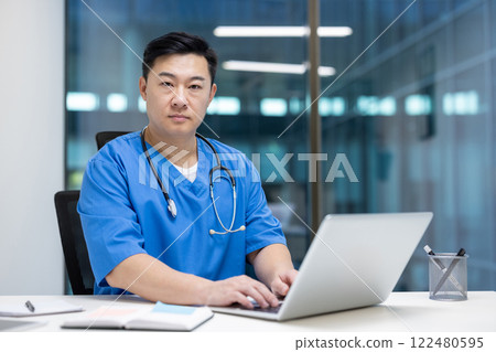Asian man doctor wearing blue scrubs, using laptop in medical office. Serious expression, highlighting focus and professionalism. Represents healthcare, technology, and dedication. Asian man doctor wearing blue scrubs, using laptop in medical office. Serious expression, highlighting focus and professionalism. Represents healthcare, technology, and dedication. 122480595