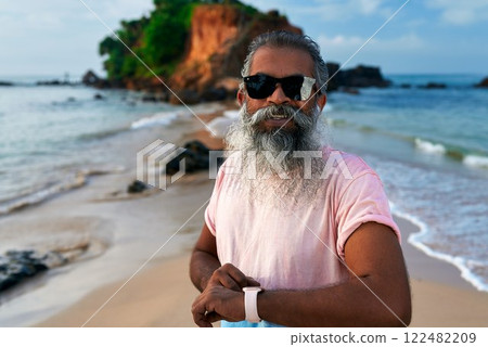 Senior South Asian man smiles on sunny beach. Wearing smart watch and earbuds. Bearded elderly using modern tech, fit lifestyle. Older adult embraces innovation, stays young at heart. Senior South Asian man smiles on sunny beach. Wearing smart watch and earbuds. Bearded elderly using modern tech, fit lifestyle. Older adult embraces innovation, stays young at heart. 122482209