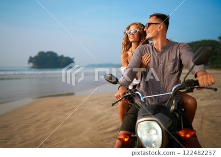 Couple on motorbike at beach. Smiling man woman enjoy getaway. Travel on budget, explore nature. Beach retreat, adventure ride. Romantic coastal journey. 122482237