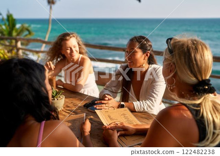 Diverse women in seaside restaurant enjoy sun. Friends choose dishes, drinks from menu. Relaxed group dining, discussing options, vacation, sunny weather, ocean view, casual atmosphere. Diverse women in seaside restaurant enjoy sun. Friends choose dishes, drinks from menu. Relaxed group dining, discussing options, vacation, sunny weather, ocean view, casual atmosphere. 122482238