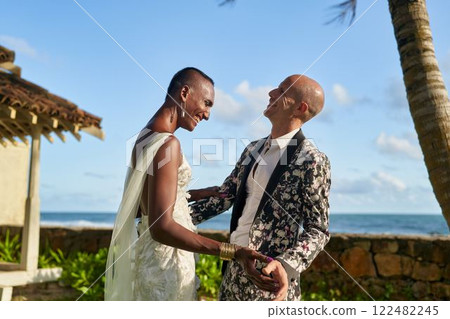 LGBTQ wedding at tropical villa. Ambiguous person in white dress as bride with partner in floral suit. Joyful couple embrace at ocean view ceremony. Celebration of love in paradise setting. 122482245