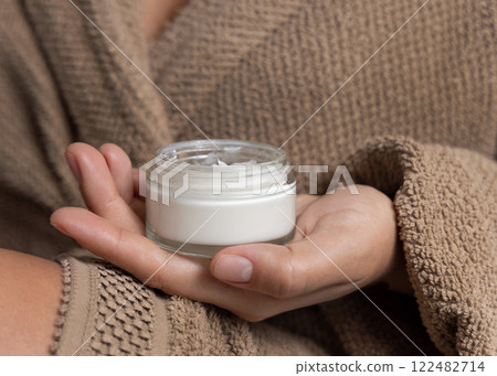 Closeup of woman in brown bathrobe holding white cream jar in hands, cosmetic mockup 122482714