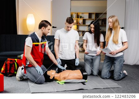 Young man instructor helping to make first aid heart compressions with dummy during the group training indoors Young man instructor helping to make first aid heart compressions with dummy during the group training indoors 122483206